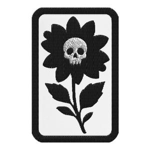 May include: Embroidered patch featuring a black flower with a skull at its center. The patch has a white background and a black border. The flower has detailed petals and leaves, and the skull is intricately designed.