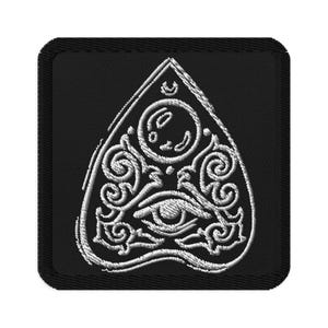 May include: Black embroidered patch featuring a white planchette design. The planchette has a crescent moon, a stylized eye, and decorative swirls. The patch has a black border and is square-shaped.
