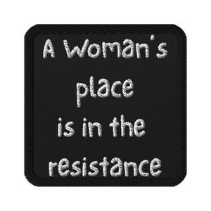 May include: Black embroidered patch with the text "A Woman's place is in the resistance" in white thread. The patch has a black border and a textured appearance, suitable for sewing or ironing onto fabric.