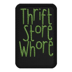 May include: Black rectangular patch with the words "Thrift Store Whore" embroidered in green, stylized lettering. The patch has a black border with a textured appearance.