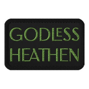 May include: A black rectangular patch with green embroidered text that reads "GODLESS HEATHEN". The patch has a stitched border and is likely designed to be sewn onto clothing or accessories.