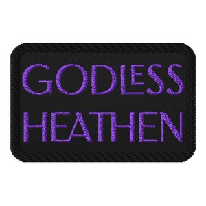 May include: Black rectangular patch with the words "GODLESS HEATHEN" embroidered in purple thread. The patch has a black border and a textured appearance.