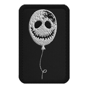 May include: Embroidered patch featuring a white balloon with a spooky face design on a black rectangular background. The balloon has two large eye holes and a jagged smile. The design is reminiscent of a Halloween theme.