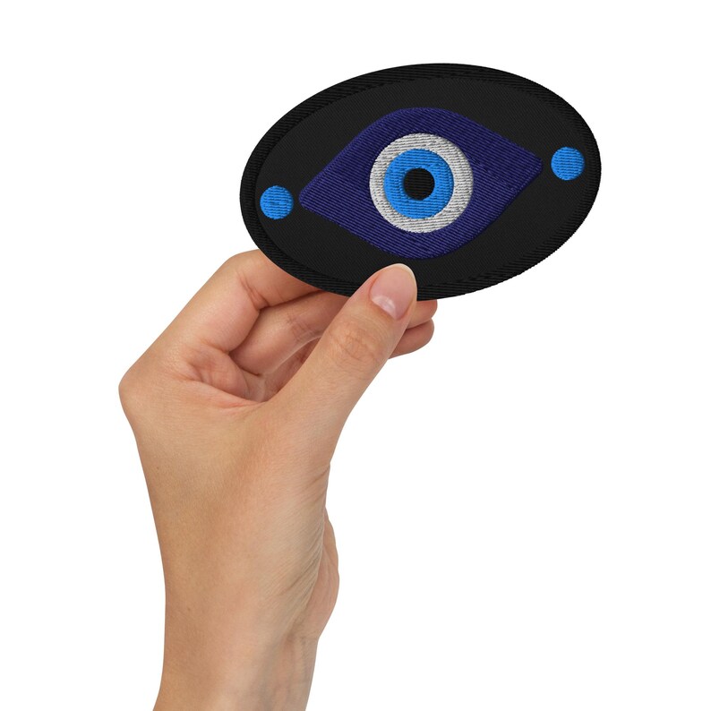 May include: Embroidered patch featuring a blue eye design on a black oval background. The eye has a blue iris, white highlights, and a black outline. Two small blue dots flank the eye. This patch is suitable for clothing or accessories.