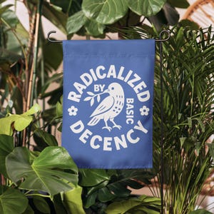 May include: A blue garden flag with white text and a dove graphic. The text reads "RADICALIZED BY BASIC DECENCY" in a circular design around the dove holding an olive branch. The flag is displayed in a garden setting.