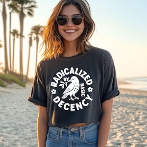 May include: A dark gray crop top with the words "RADICALIZED BY BASIC DECENCY" in a white circular design. The design includes a dove holding an olive branch. The model is wearing blue jeans and sunglasses.