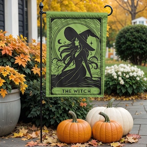 May include: A rectangular garden flag featuring a black graphic illustration of a witch wearing a pointed hat against a light green background. The design includes a crescent moon, stars, and swirling clouds. The text THE WITCH is printed at the bottom.