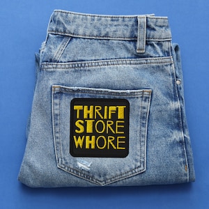 May include: Light blue denim jeans with a black patch on the back pocket. The patch features the words "THRIFT STORE WHORE" in yellow, embroidered lettering. The jeans are folded and set against a blue background.