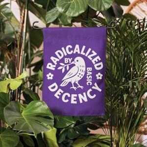 May include: A purple garden flag with the words "RADICALIZED BY BASIC DECENCY" in white, surrounding a white dove holding an olive branch. The flag is displayed outdoors among green plants.