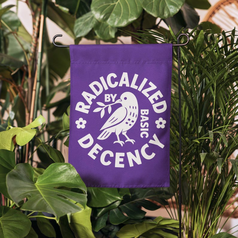 May include: A purple garden flag with the words "RADICALIZED BY BASIC DECENCY" in white, surrounding a white dove holding an olive branch. The flag is displayed outdoors among green plants.