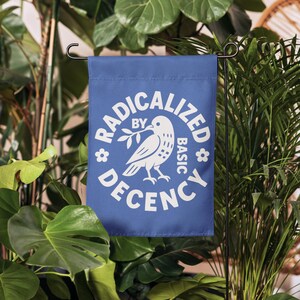 May include: A blue garden flag with white text and a dove graphic. The text reads "RADICALIZED BY BASIC DECENCY". The dove holds an olive branch. The flag is displayed outdoors among green plants.