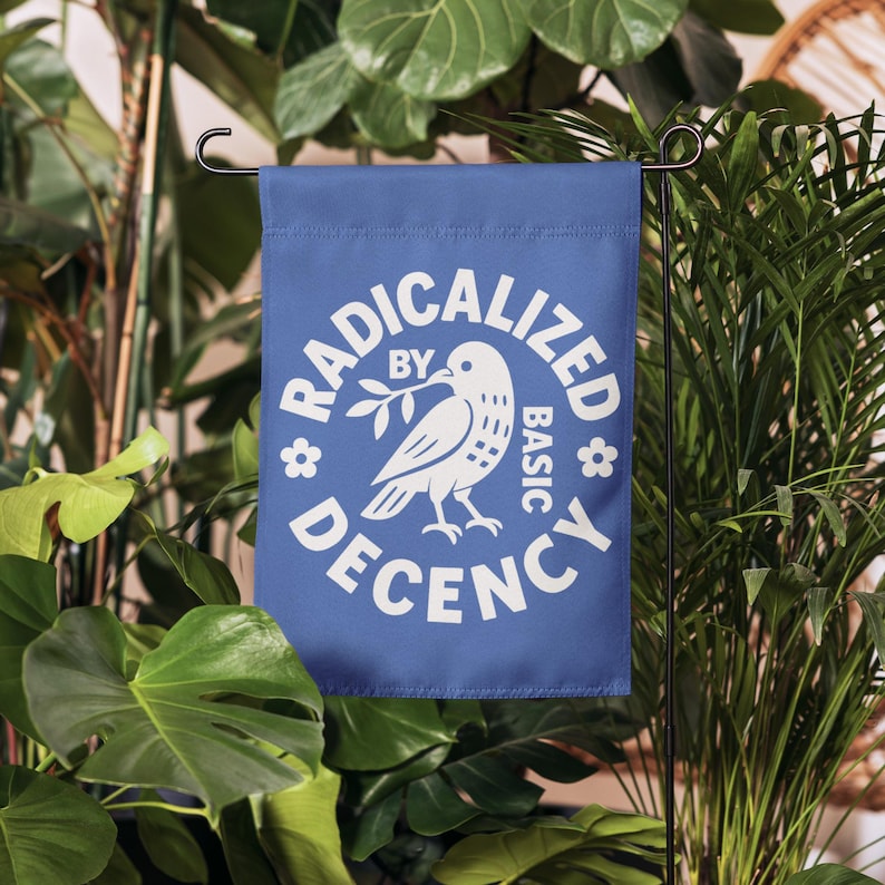 May include: A blue garden flag with white text and a dove graphic. The text reads "RADICALIZED BY BASIC DECENCY". The dove holds an olive branch. The flag is displayed outdoors among green plants.