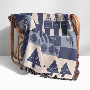 May include: A blue and white woven throw blanket with a Christmas theme. The blanket features a pattern of nutcrackers, ornaments, and Christmas trees. The blanket has a fringe border.
