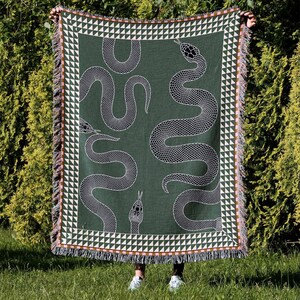 Puede incluir: Una manta tejida verde con un patr&oacute;n de serpiente blanca. La manta tiene un borde de flecos con un patr&oacute;n geom&eacute;trico.