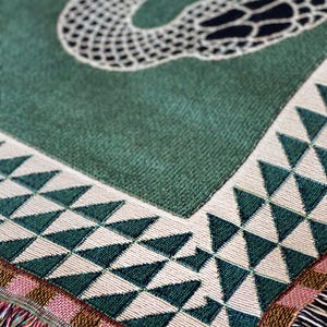 Puede incluir: Una manta de tejido verde con un patr&oacute;n geom&eacute;trico de tri&aacute;ngulos blancos y verdes. La manta tiene un borde de flecos.
