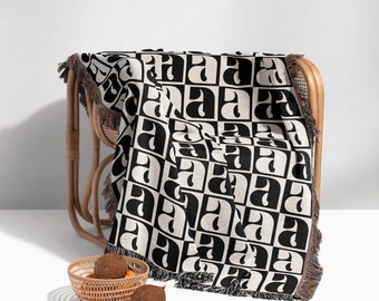 May include: Black and white woven throw blanket with a geometric pattern of the letter 'A' repeated. The blanket is draped over a wicker chair with a fringe border.