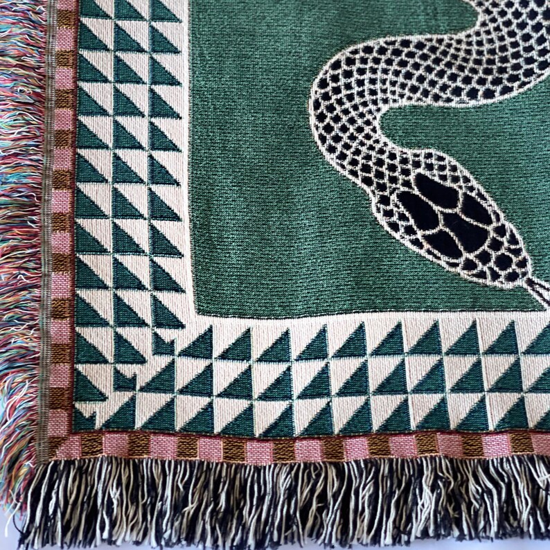 May include: A green woven throw blanket featuring a black snake design. The blanket has a fringe border with pink, brown and white stripes.