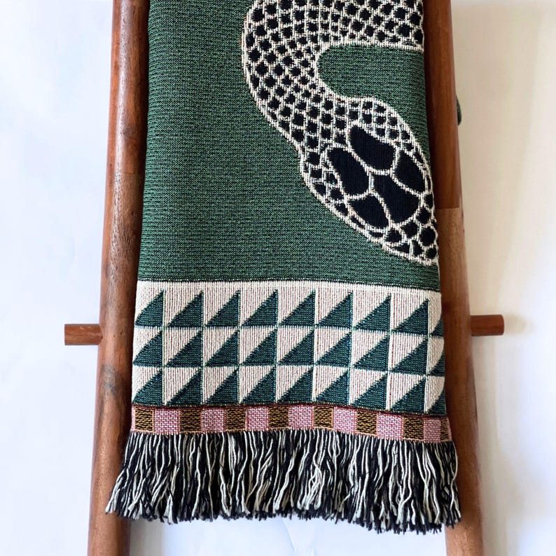 Green woven cotton throw blanket draped on a wooden ladder, featuring an off white-and-black stylized snake motif with a border of white triangular geometric patterns and finished with multicolored fringe