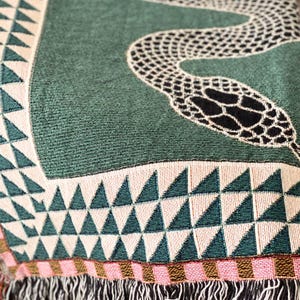 May include: A green woven throw blanket featuring a black snake design and a geometric pattern of white and green triangles. The blanket has a fringed border.