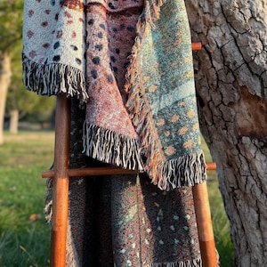 May include: A collection of woven throw blankets in various colors, including teal, burgundy, and cream, with fringed edges. The blankets are draped over a wooden ladder, showcasing their different patterns and textures. The patterns include polka dots and floral designs.
