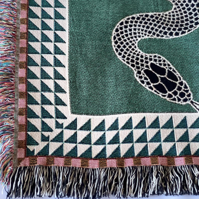 Puede incluir: Una manta tejida verde con un dise&ntilde;o de serpiente negra. La manta tiene un borde de flecos con rayas rosas, marrones y blancas.
