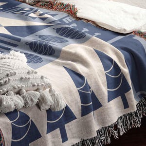 May include: A woven throw blanket with a cream background and a blue geometric pattern of stylized trees. The blanket has fringe on all sides.