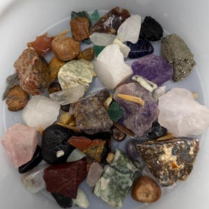 May include: A collection of various raw gemstones and crystals in a white container. The assortment includes amethyst, rose quartz, clear quartz, and other colorful stones. The stones vary in size and shape, with a mix of textures and colors.