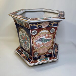May include: An octagonal ceramic planter with a matching saucer. The planter features a dark blue, burgundy, and gold color scheme with floral and bird motifs. The design is reminiscent of traditional Asian art.