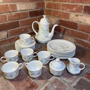 May include: A white porcelain tea set with a floral pattern in blue and green. The set includes a teapot, cups, saucers, plates, a sugar bowl, and a creamer. The tea set is arranged on a brick surface.