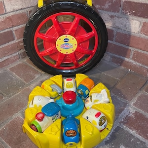 May include: A yellow and black toy tire with a red center and a yellow handle. Below it is a yellow toy with various colorful vehicles and a red button in the center. The toys are designed for children.