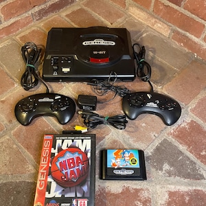 May include: A black Sega Genesis 16-bit video game console with two controllers, power adapter, and two game cartridges. One game cartridge is "NBA Jam" and the other is "Sonic the Hedgehog". The console and accessories are on a brick surface.