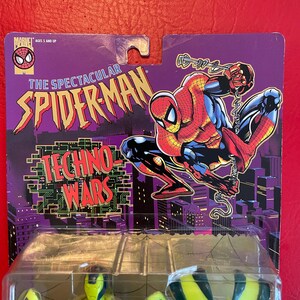 May include: A yellow and green action figure of Spider-Man in his Techno-Wars suit. The figure is packaged in a plastic blister pack with a purple and black card featuring a cartoon image of Spider-Man. The card reads "The Spectacular Spider-Man Techno-Wars".