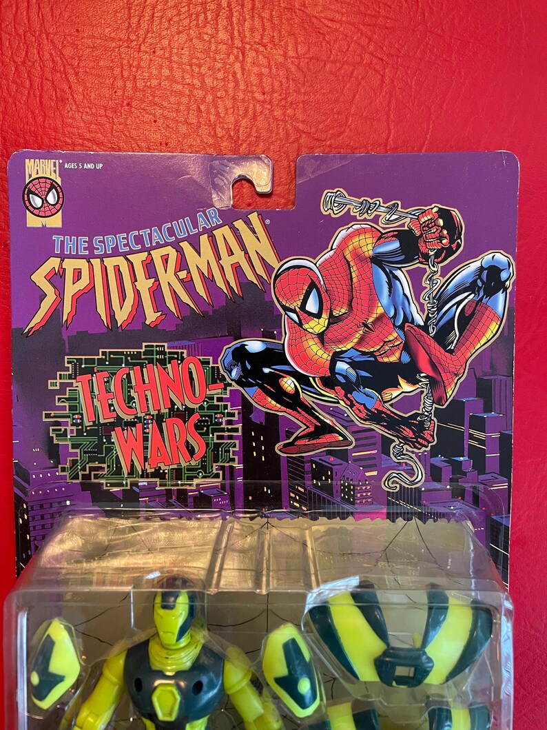 May include: A yellow and green action figure of Spider-Man in his Techno-Wars suit. The figure is packaged in a plastic blister pack with a purple and black card featuring a cartoon image of Spider-Man. The card reads "The Spectacular Spider-Man Techno-Wars".