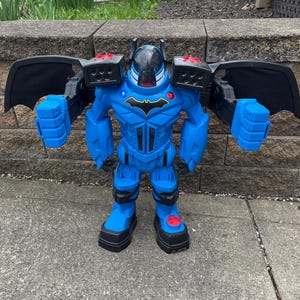 May include: A blue Batman action figure with black wings, red accents, and a Batman logo on the chest. The toy robot has a clear helmet and large blue fists. The figure is standing on a concrete surface.