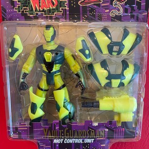 May include: A green and yellow action figure of a robot with a black chest plate. The figure is holding a yellow gun and comes with additional armor pieces. The packaging features the text "Techno Wars" and "Vault Guardsman Riot Control Unit".