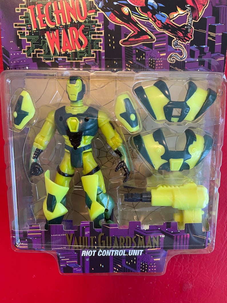 May include: A green and yellow action figure of a robot with a black chest plate. The figure is holding a yellow gun and comes with additional armor pieces. The packaging features the text "Techno Wars" and "Vault Guardsman Riot Control Unit".