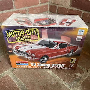 May include: A Monogram model kit box featuring a red '66 Shelby GT350 with white racing stripes. The box reads "MOTOR-CITY MUSCLE" and includes the text "1 PLASTIC KIT" and "1:24". The box is on a brick surface.