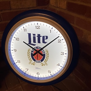 May include: A white and blue round wall clock with a blue outer ring and a white face. The clock features the Lite beer logo with the text "A Fine Pilsner Beer". The clock hands are black and the numbers are black and white.