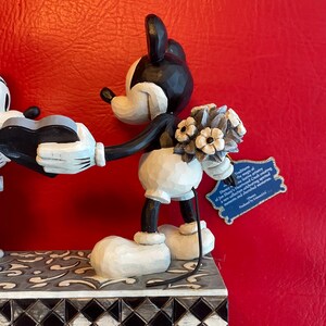 Pu&ograve; includere: Figurina in bianco e nero di Topolino che tiene un mazzo di fiori e un cuore. La figurina &egrave; in piedi su una base a scacchi bianchi e neri con un motivo floreale grigio.