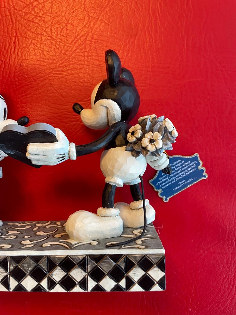 Pu&ograve; includere: Figurina in bianco e nero di Topolino che tiene un mazzo di fiori e un cuore. La figurina &egrave; in piedi su una base a scacchi bianchi e neri con un motivo floreale grigio.