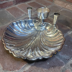 May include: A silver-colored, shell-shaped serving dish with a central bowl and two small, cylindrical holders. The dish has a scalloped edge and a reflective surface, likely made of metal. The design is ornate and decorative.