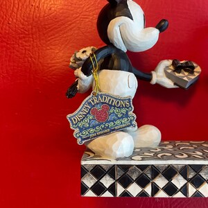 Pu&ograve; includere: Una statuina di Topolino in bianco e nero che tiene un mazzo di fiori e una scatola a forma di cuore. La statuina &egrave; in piedi su una base a scacchi bianchi e neri con un'etichetta Disney Traditions Jim Shore Enesco.