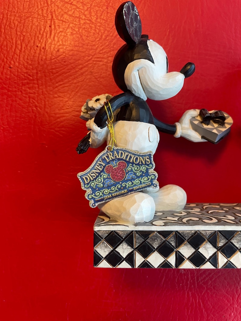 Pu&ograve; includere: Una statuina di Topolino in bianco e nero che tiene un mazzo di fiori e una scatola a forma di cuore. La statuina &egrave; in piedi su una base a scacchi bianchi e neri con un'etichetta Disney Traditions Jim Shore Enesco.