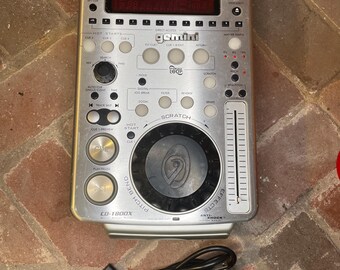May include: Silver Gemini CD-1800X DJ CD player with various buttons and a central scratch wheel. Features a digital display and a black power cord. The unit is on a brick surface.