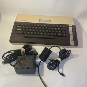 May include: An Atari 800XL computer with a black keyboard and beige casing. The image includes a power supply and cables. The keyboard has black keys with white lettering. The Atari 800XL is a vintage computer.