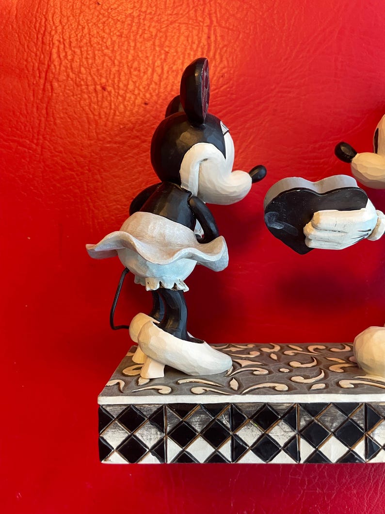 Pu&ograve; includere: Una statuetta in bianco e nero di Topolino e Minnie Mouse che tiene un cuore. La statuetta &egrave; su una base piastrellata in bianco e nero.