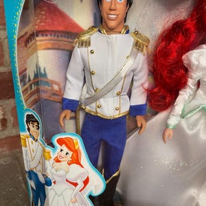 May include: A Prince Eric doll from Disney's The Little Mermaid, in a clear plastic package. The doll wears a white jacket with gold trim, blue pants, and black boots. The packaging features illustrations of Ariel and Eric.