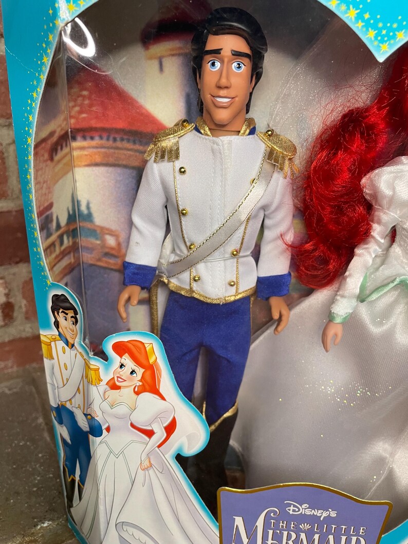 May include: A Prince Eric doll from Disney's The Little Mermaid, in a clear plastic package. The doll wears a white jacket with gold trim, blue pants, and black boots. The packaging features illustrations of Ariel and Eric.