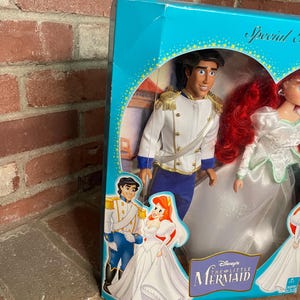 May include: A boxed set of Disney's The Little Mermaid dolls. The dolls are Prince Eric and Ariel in wedding attire. The box is teal with gold accents and features illustrations of the characters. The box reads "Special Edition".