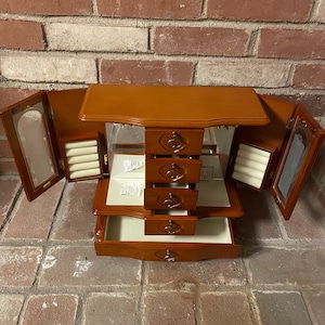 May include: A wooden jewelry box with a mirrored interior and multiple drawers. The box has a heart-shaped design on the front and is decorated with a gold-colored knob.